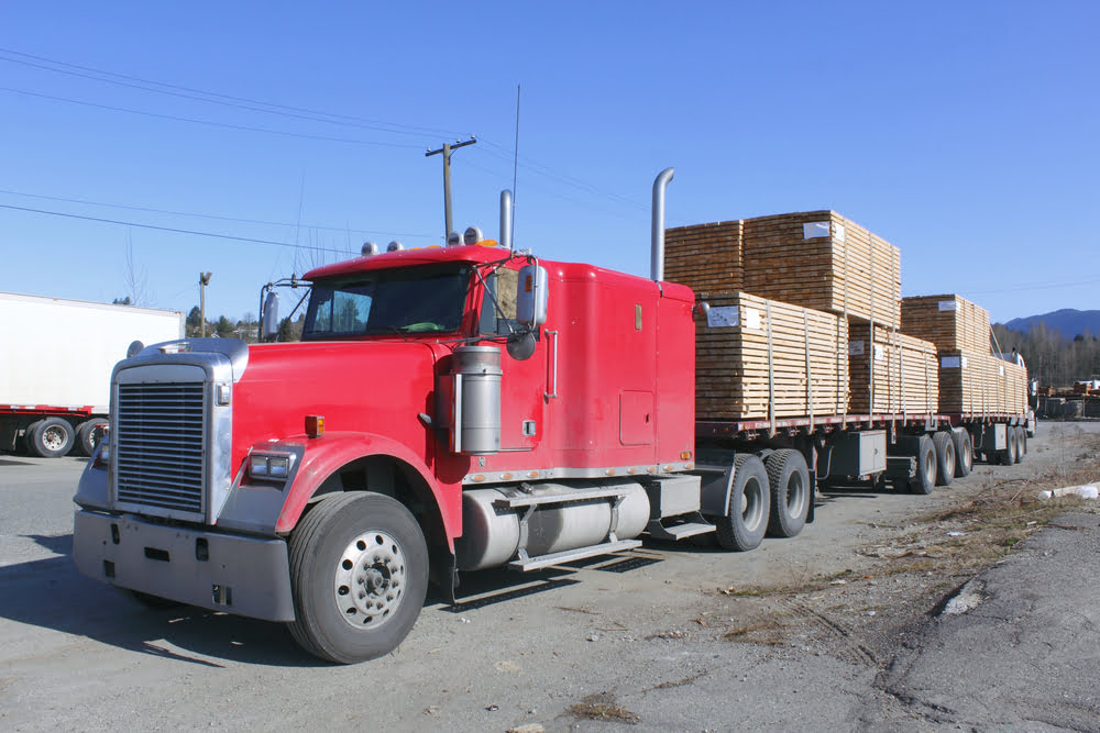 Red heavy hauling truck with stacked lumber secured on flatbed trailers.